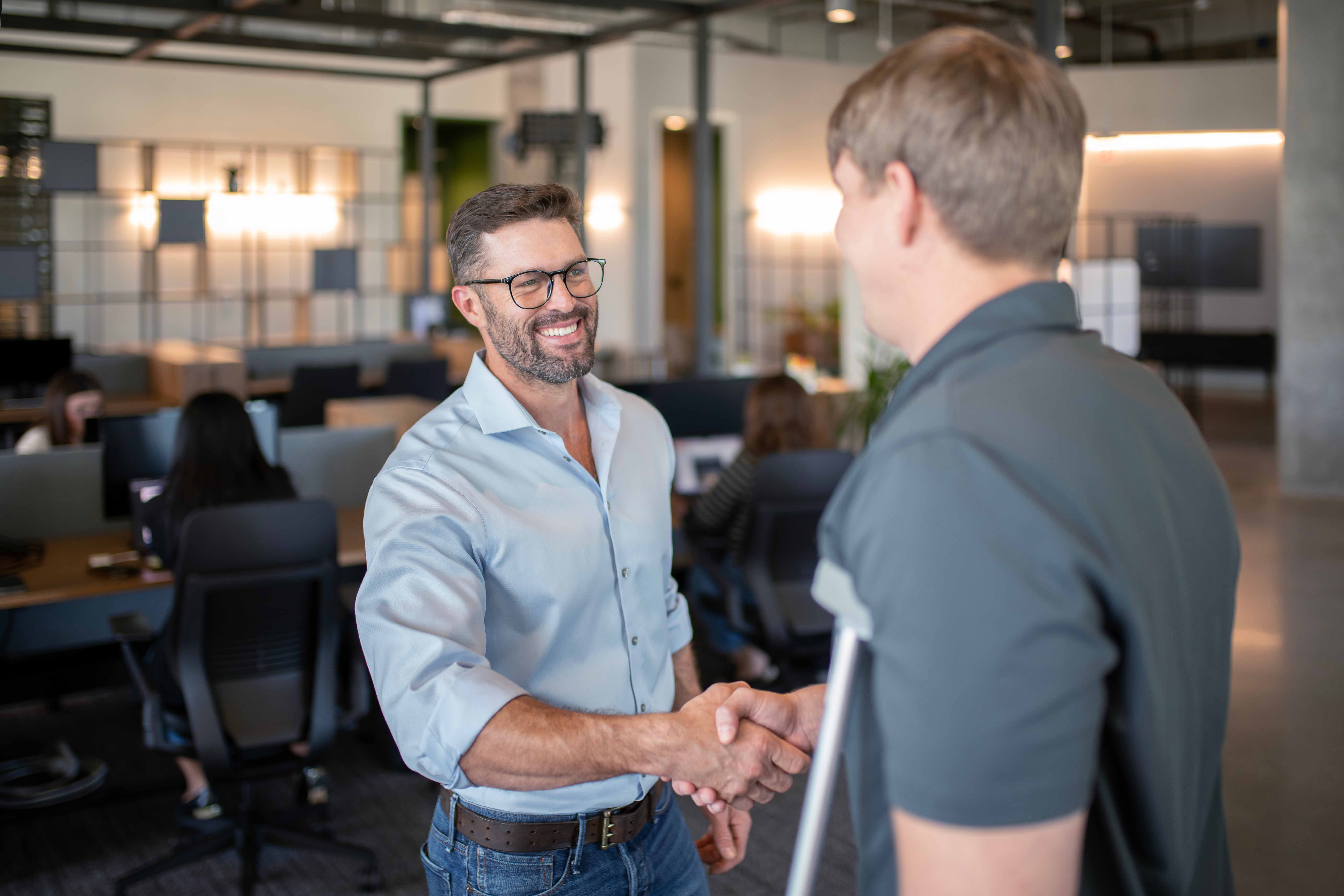 A man in a light blue shirt smiles while shaking hands with another person using crutches, standing in an office environment with other employees working at desks in the background.